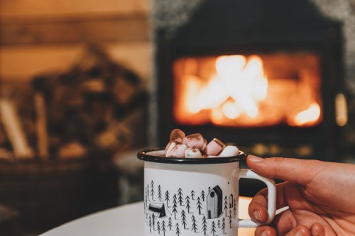 A hand holds a mug of hot chocolate topped with marshmallows, with a cozy fireplace in the background.
