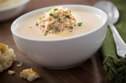 A bowl of creamy soup topped with shredded chicken and parsley, with a piece of bread nearby.