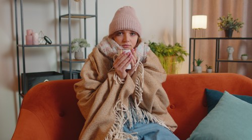 A person wrapped in a cozy blanket and scarf, holding a mug, sitting on a couch in a warmly decorated room.
