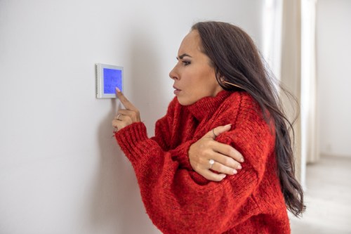 A woman in a red sweater looks frustrated while adjusting a thermostat on the wall.