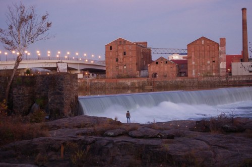 A person stands near a waterfall, with old industrial buildings and a bridge in the background at dusk.
