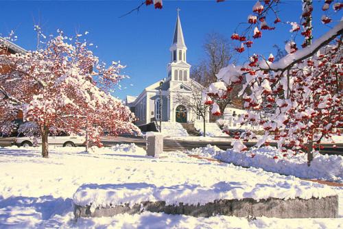 A snowy scene featuring a church with a steeple, surrounded by trees with red leaves and a clear blue sky.