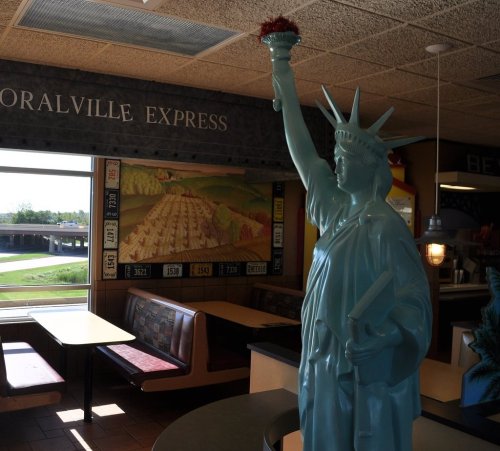 A teal statue of the Statue of Liberty stands inside a restaurant, with a mural and seating in the background.