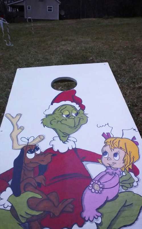 A festive cornhole board featuring the Grinch with a reindeer and a girl, all in holiday attire.