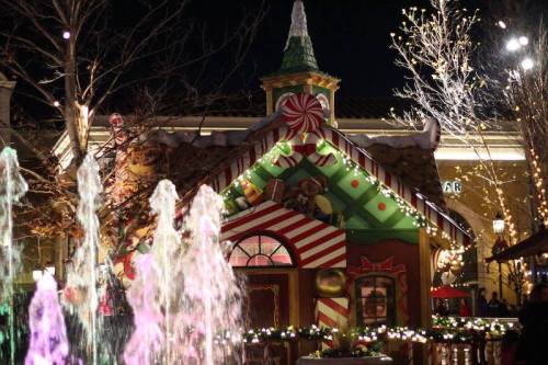 A festive gingerbread house decorated with candy canes, surrounded by colorful lights and a fountain at night.
