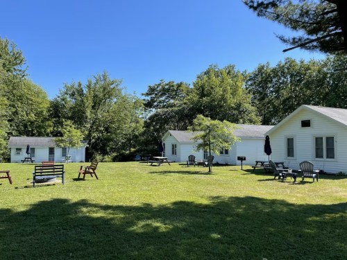 A sunny outdoor scene featuring two white cabins surrounded by green grass and trees, with benches and picnic tables.