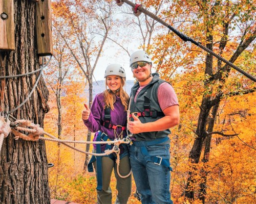 Two people in helmets give thumbs up while standing on a platform surrounded by colorful autumn trees.