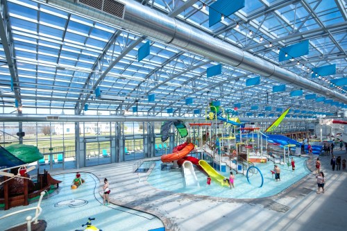 Bright indoor water park featuring colorful slides, play structures, and families enjoying the water. Large glass roof above.