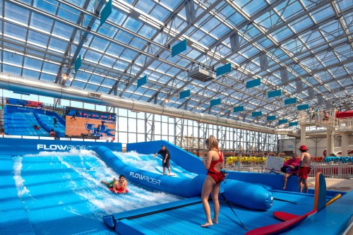 A vibrant indoor water park scene featuring a FlowRider surf simulator with people surfing and lifeguards on duty.
