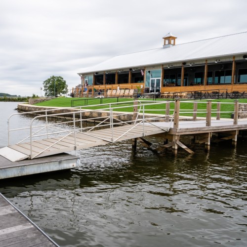 A wooden dock leads to a restaurant by a calm lake, with green grass and a cloudy sky in the background.