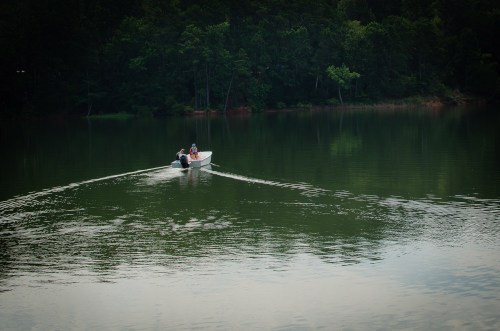 A small boat with two people glides across a calm, green lake surrounded by trees.