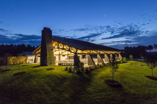 A modern building with a stone chimney, illuminated at dusk, surrounded by landscaped greenery and a cloudy sky.