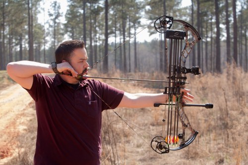 A person in a maroon shirt aims a compound bow in a wooded area, preparing to shoot an arrow.