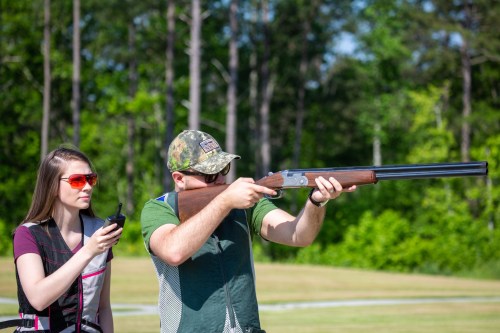 A person aims a shotgun while another observes with a walkie-talkie, set in a green outdoor shooting range.