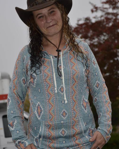 A person with wet, curly hair wearing a patterned hoodie and a cowboy hat, smiling outdoors.