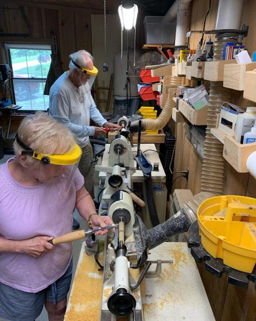 Two people working on a wood lathe in a workshop, wearing safety goggles and using tools amidst wood shavings.