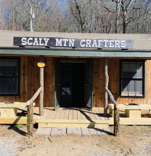 A rustic wooden building with a sign reading "Scaly Mtn Crafters" and a porch, surrounded by trees.