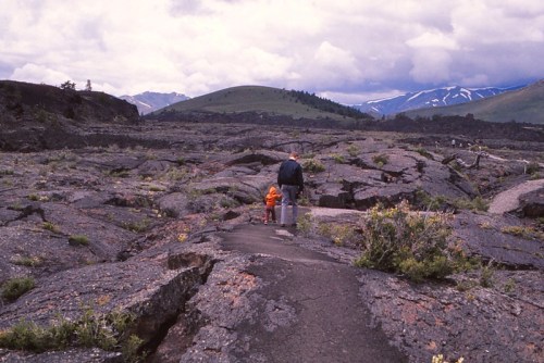 A person and a child walk on rocky terrain with mountains in the background under a cloudy sky.