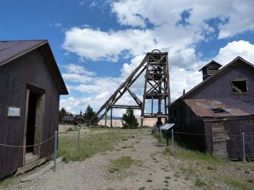 Abandoned mining structures with a tall headframe against a blue sky and scattered clouds.