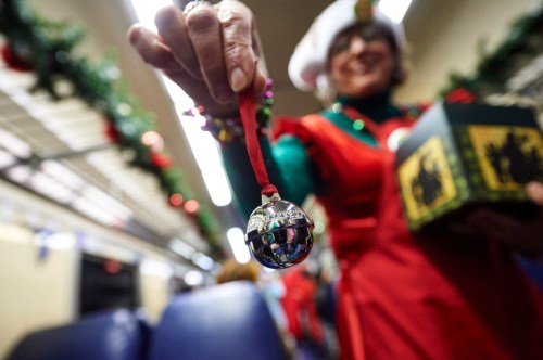 A person in a festive outfit holds a jingle bell and a gift box, surrounded by holiday decorations on a train.