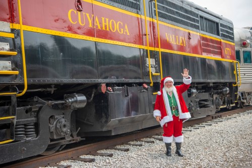 Santa Claus waves beside a vintage train in Cuyahoga Valley, dressed in his traditional red suit and hat.