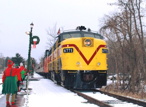 A bright yellow train stops at a snowy station, with festive elves waving nearby and holiday decorations adorning the scene.