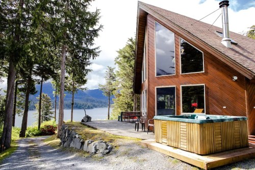 A modern cabin with large windows, a hot tub, and a scenic view of trees and water in the background.