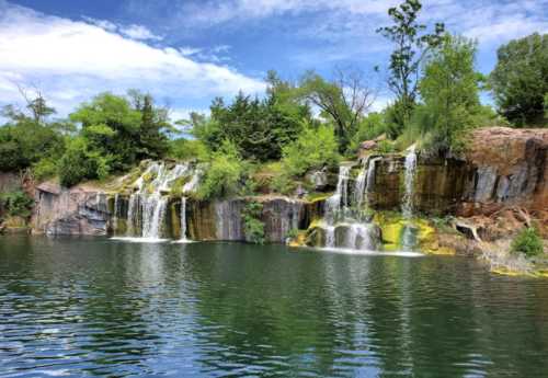 A serene waterfall cascading over rocky cliffs into a calm, green-tinted lake surrounded by lush trees and blue sky.