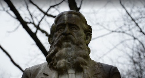 A close-up of a weathered statue of a bearded man in a suit, surrounded by bare tree branches.