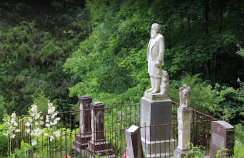 A statue of a man stands in a green cemetery, surrounded by old gravestones and a wrought iron fence.