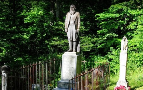 A tall statue of a man stands in a green forest, surrounded by a wrought iron fence and a stone monument.