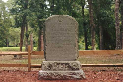 A weathered stone monument stands in a wooded area, with inscriptions and the year 1896 visible at the base.