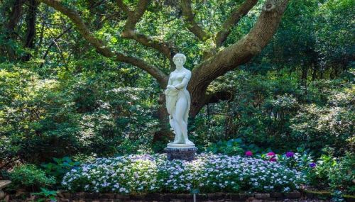 A white statue of a woman stands amidst vibrant flowers and lush greenery under a large tree.