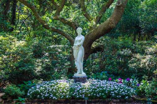 A white statue of a woman stands amidst vibrant flowers and lush greenery in a serene garden setting.