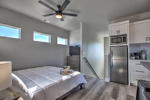 Cozy bedroom with a bed, ceiling fan, TV, and kitchenette, featuring large windows and modern decor.