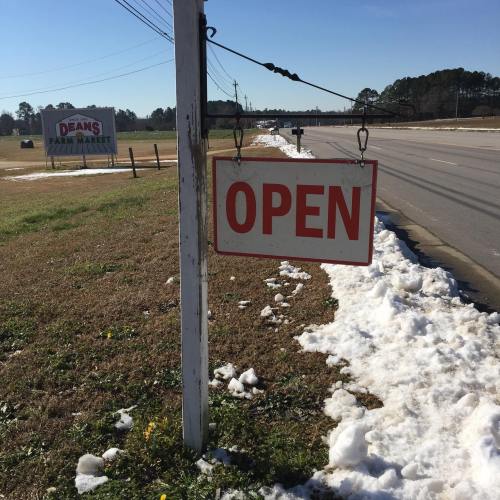 A roadside sign with the word "OPEN" hanging, surrounded by snow and grass, near a road.