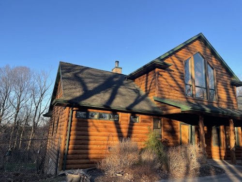 A log cabin with large windows, surrounded by trees and a clear blue sky. Shadows cast on the exterior.