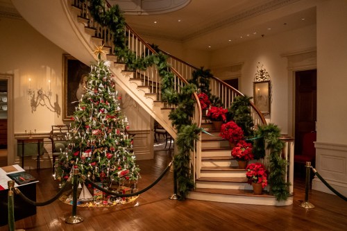 A beautifully decorated Christmas tree and festive garlands adorn a grand staircase in a warmly lit interior.