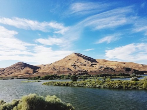 A serene landscape featuring a river, lush greenery, and rolling hills under a blue sky with scattered clouds.