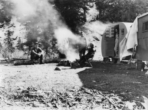 A black-and-white photo of campers by a fire, with two trailers and trees in the background. Smoke rises from the fire.