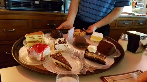 A person holding a tray with various desserts, including cakes and pastries, in a cozy restaurant setting.