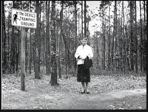 A woman stands near a sign reading "The Devil's Tramping Ground" in a wooded area, surrounded by trees.