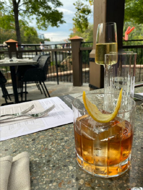 A cocktail with a lemon twist sits on a table beside a glass of champagne, with a menu and outdoor scenery in the background.