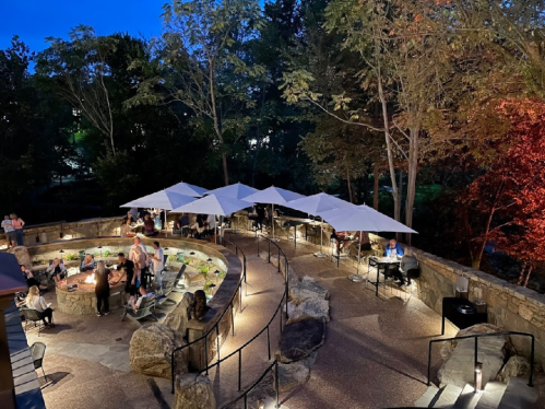 A cozy outdoor setting at dusk with umbrellas, seating areas, and people enjoying the ambiance near a stone pathway.