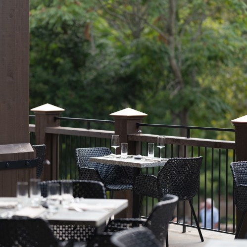 Outdoor dining area with empty tables and chairs, surrounded by greenery and a wooden railing.