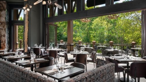 Modern restaurant interior with wooden accents, elegant tables, and large windows overlooking a lush green landscape.