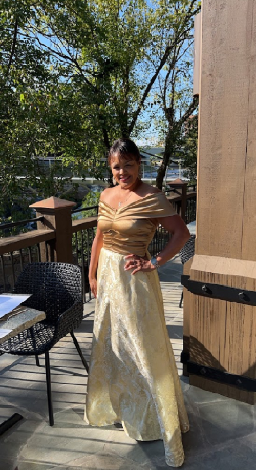 A woman poses outdoors in a stylish off-shoulder top and a long, elegant skirt, surrounded by trees and a patio setting.