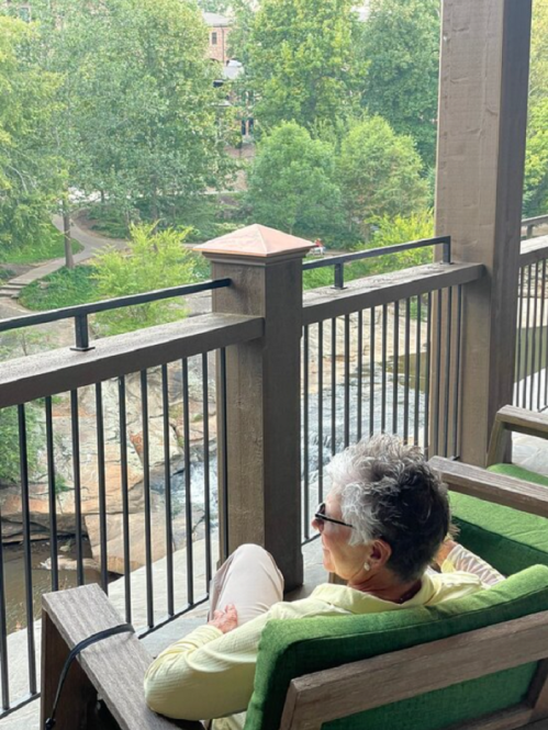An older woman sits on a balcony, gazing at a lush green landscape and a waterfall below.