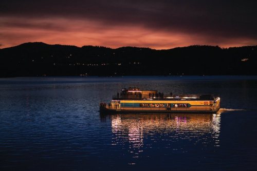 A boat illuminated at dusk glides across a calm lake, surrounded by dark hills and a colorful sunset sky.