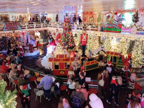 A festive holiday scene with decorated trees, lights, a train, and people enjoying a Christmas-themed event.